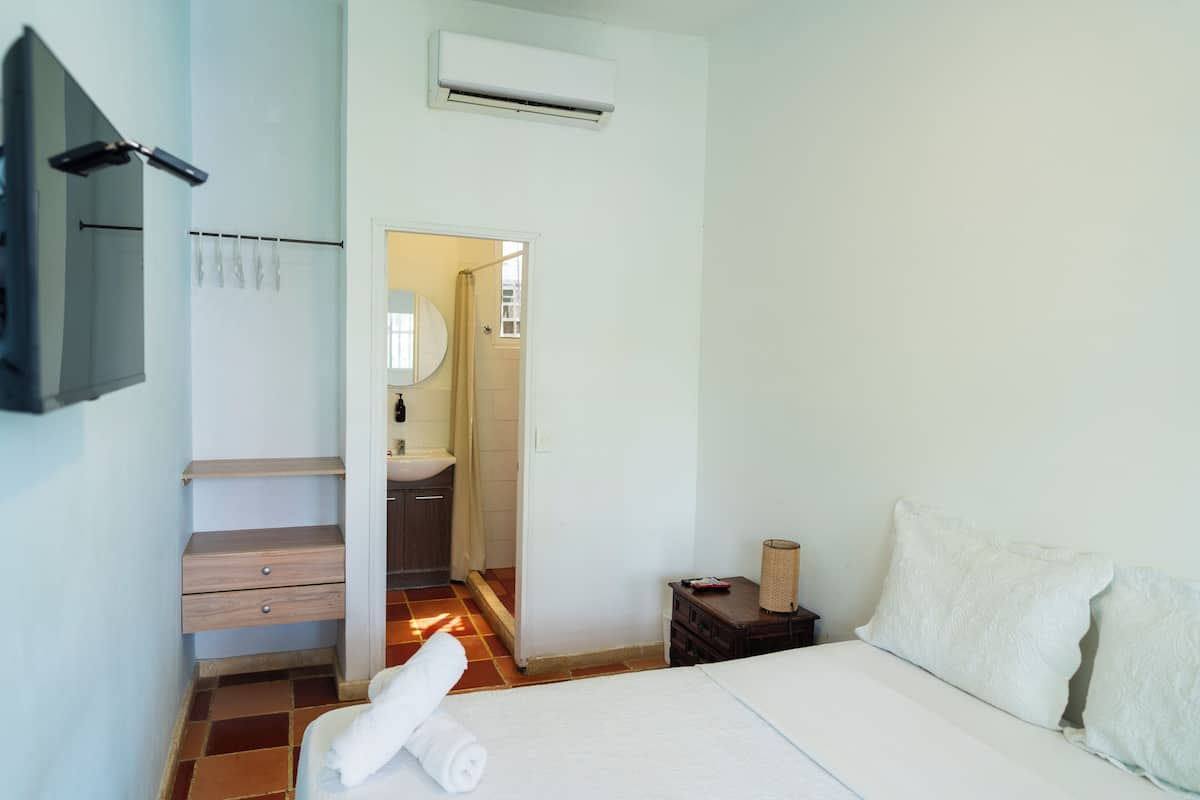 A bedroom with a bed, a bedside table, a nightstand, a wall-mounted air conditioner, a mirror, and a bathroom visible through an open door.