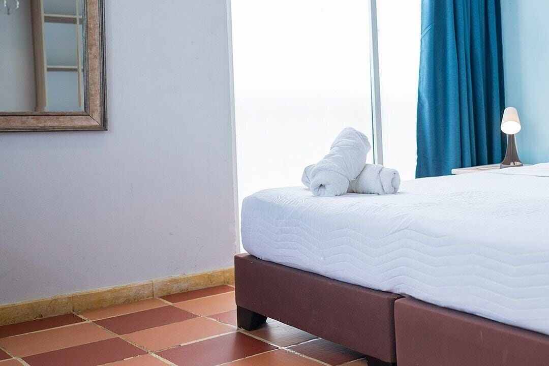 Une chambre d’hôtel avec un lit, une table de chevet, une lampe, une fenêtre avec des rideaux et un miroir au mur.