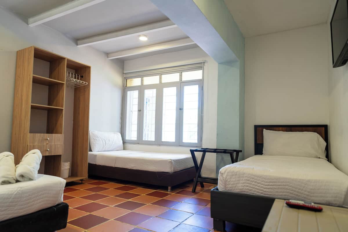A room with two beds, a wooden wardrobe, a small table, and a window with blinds.