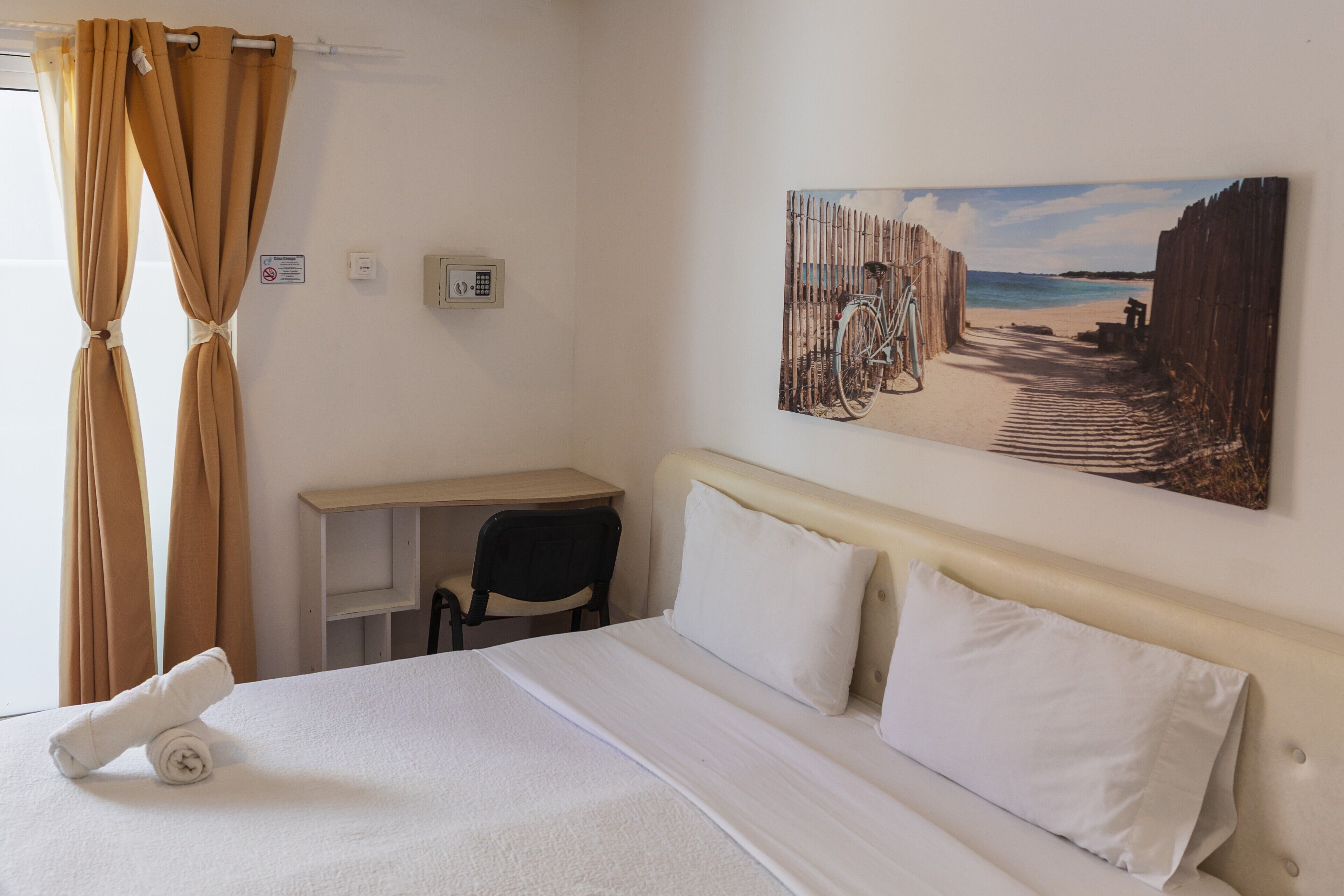A hotel room with a bed, a desk, a chair, a painting on the wall, and curtains.
