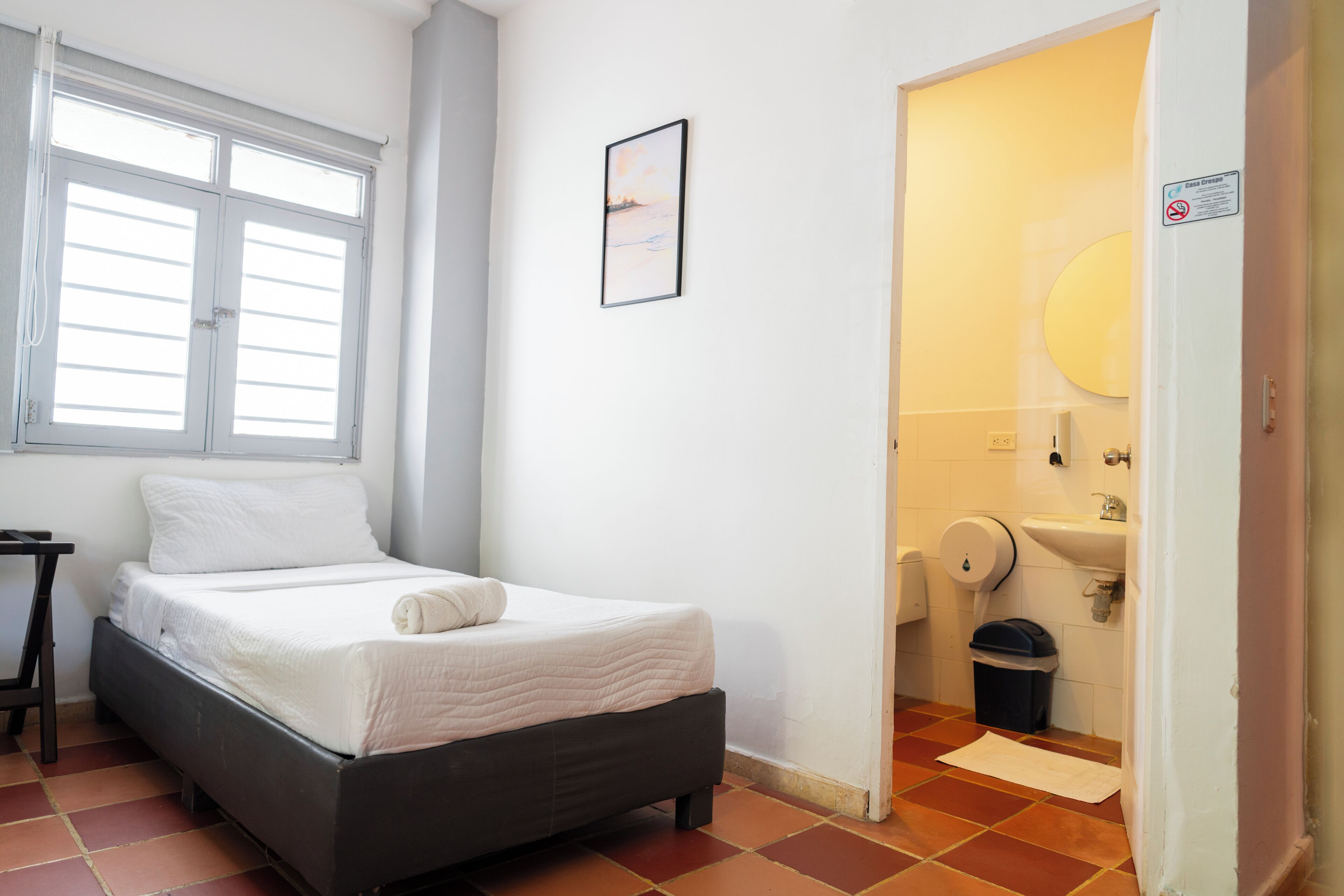 Un dormitorio con cama, ventana, puerta que da a un baño y una mesita.