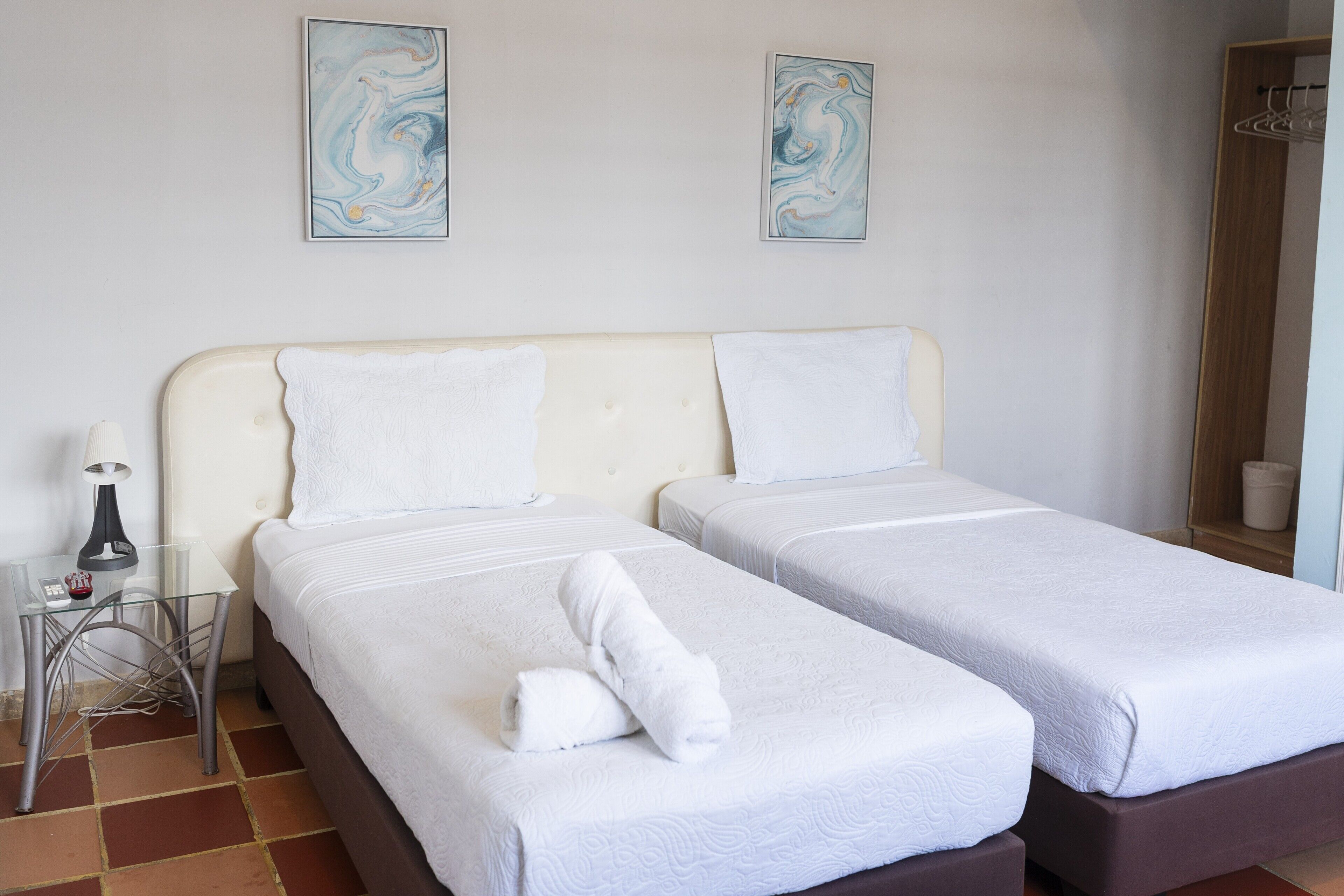 A hotel room with two beds, a bedside table with a lamp, and abstract wall art.
