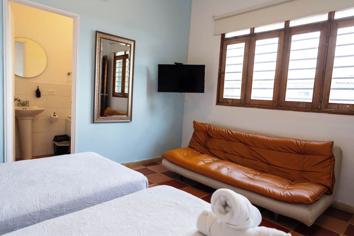 A hotel room with two beds, a sofa, a TV, a mirror, and a window with blinds.