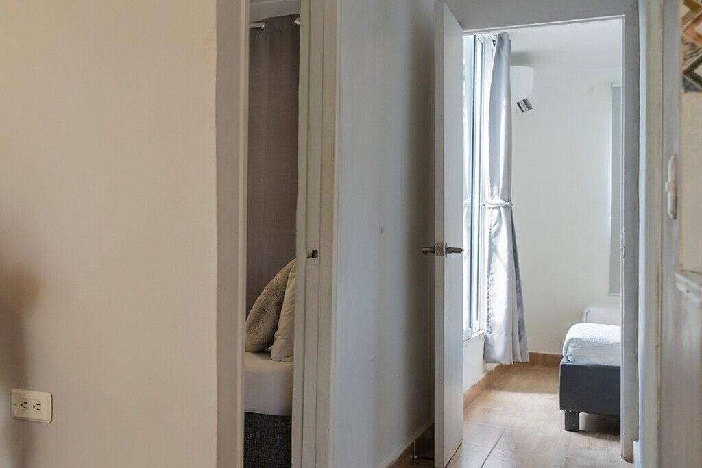 A hallway leading to a bedroom with a bed, a nightstand, and a window with curtains.