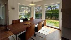 Une salle à manger moderne avec une table et des chaises en bois, de grandes fenêtres donnant sur un jardin et des maisons, et un luminaire au plafond.