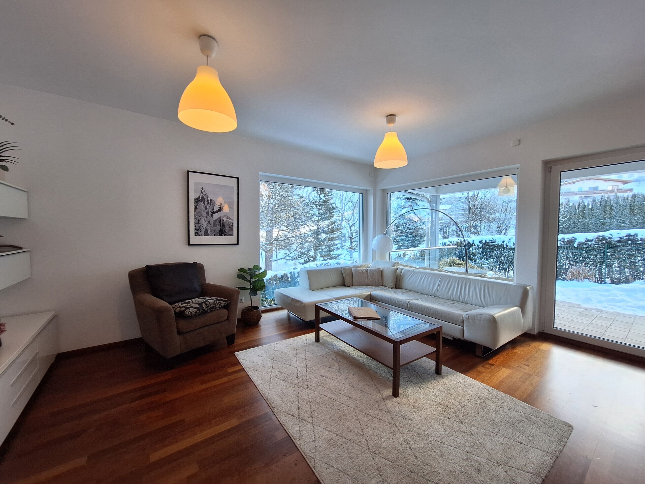 Una sala de estar moderna con sofá, sillón y mesa de centro. Amplias ventanas ofrecen vistas a un paisaje nevado.