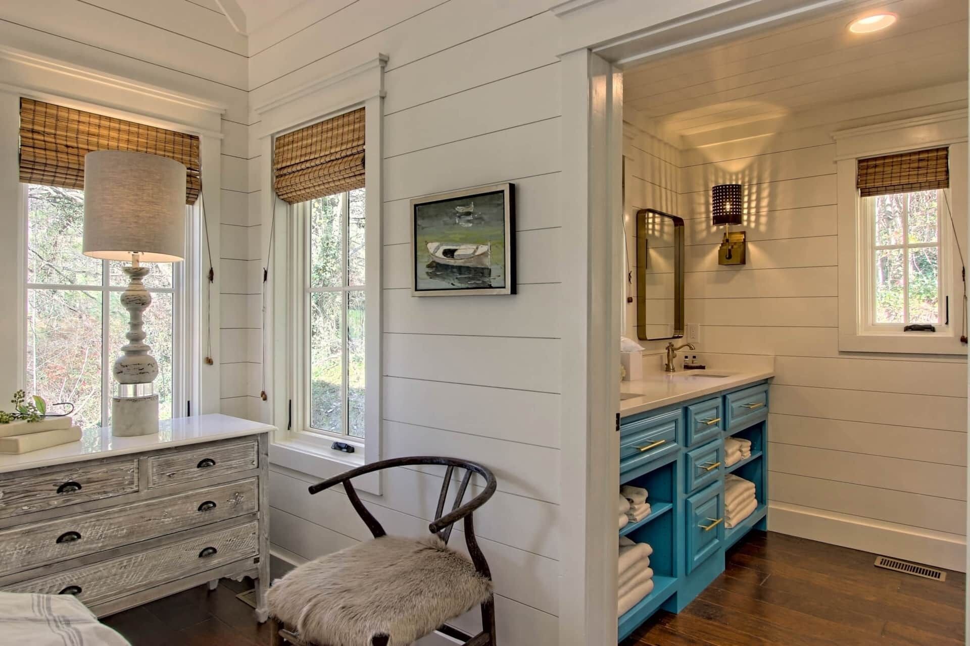 A bathroom with white paneled walls, a blue vanity, a chair, and a framed picture on the wall.