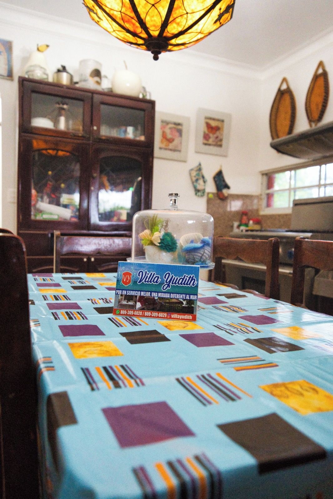 Uma mesa de jantar com toalha de mesa colorida, um recipiente de vidro com objetos decorativos e uma placa com o nome Villa Yudith.