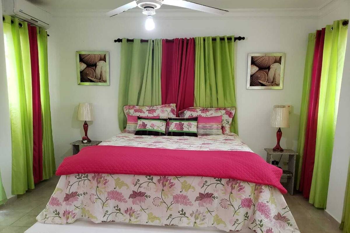 Un dormitorio con una cama, dos mesitas de noche, un ventilador de techo y cortinas.