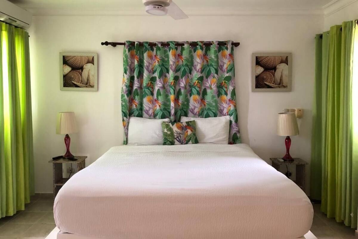 Una habitación de hotel con cama, cortinas verdes, un ventilador de techo y dos lámparas de noche.