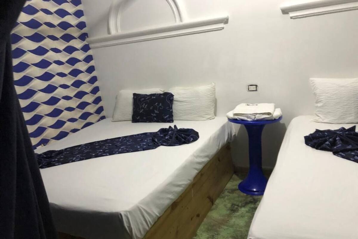 Una habitación pequeña y funcional con dos camas individuales, una mesita y una cortina estampada.