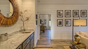 A modern kitchen with a large mirror, a sink, and a countertop. There is a hallway leading to a living area with a sofa and a lamp. - Sweet Tea Cottage at Julep Farms (Dillard)