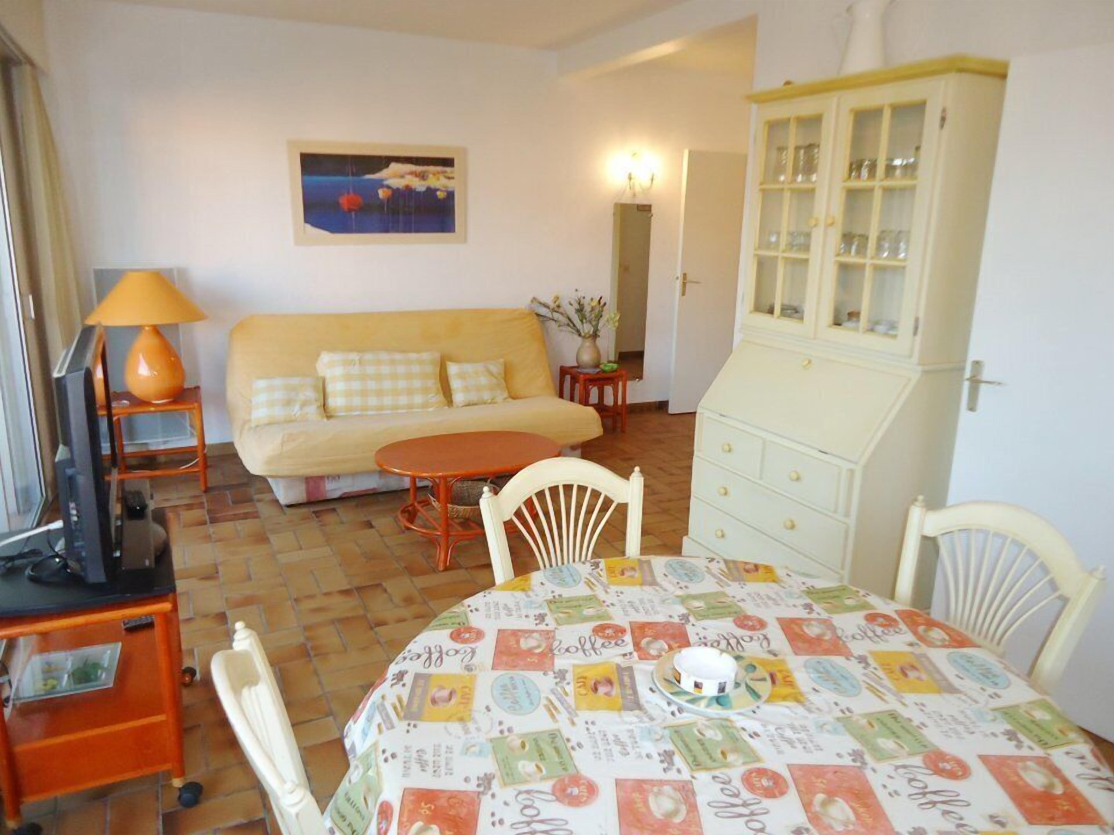 A cozy living room with a sofa, a dining table with a coffee-themed tablecloth, a cabinet, and a television.