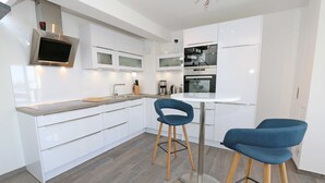 Una cocina moderna con armarios blancos, horno empotrado, microondas y cafetera. Hay dos taburetes azules y una mesa redonda.