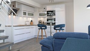 Una cocina moderna con armarios blancos, horno integrado y cafetera. En la zona de estar, hay dos taburetes azules y un sofá azul.