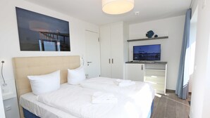 Una cama bien hecha con sábanas blancas, un televisor sobre un mueble bajo y un cuadro de un puente en la pared.