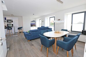 Un salon moderne avec un canapé bleu, une table à manger avec des chaises et un coin cuisine doté de meubles blancs et d’un téléviseur.