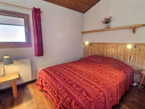 A bedroom with a wooden headboard, a bed with a patterned bedspread, a nightstand with a lamp, a window with curtains, and a shelf with a vase.