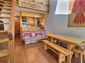 A cozy room with a wooden bench, a sofa with patterned cushions, a wooden table, and a kitchen area with wooden cabinets.