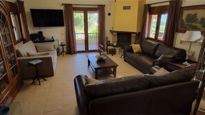 Ein Wohnzimmer mit Sofa, Sessel, Couchtisch und Kamin. Ein Fernseher ist an der Wand montiert und es gibt ein Fenster mit Vorhängen.