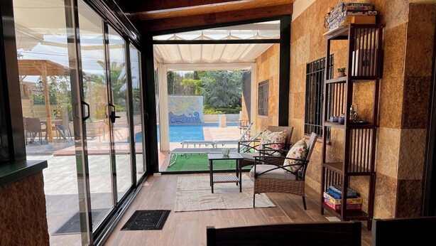 A modern room with a glass door leading to a pool area, a wicker chair, and a bookshelf with board games.
