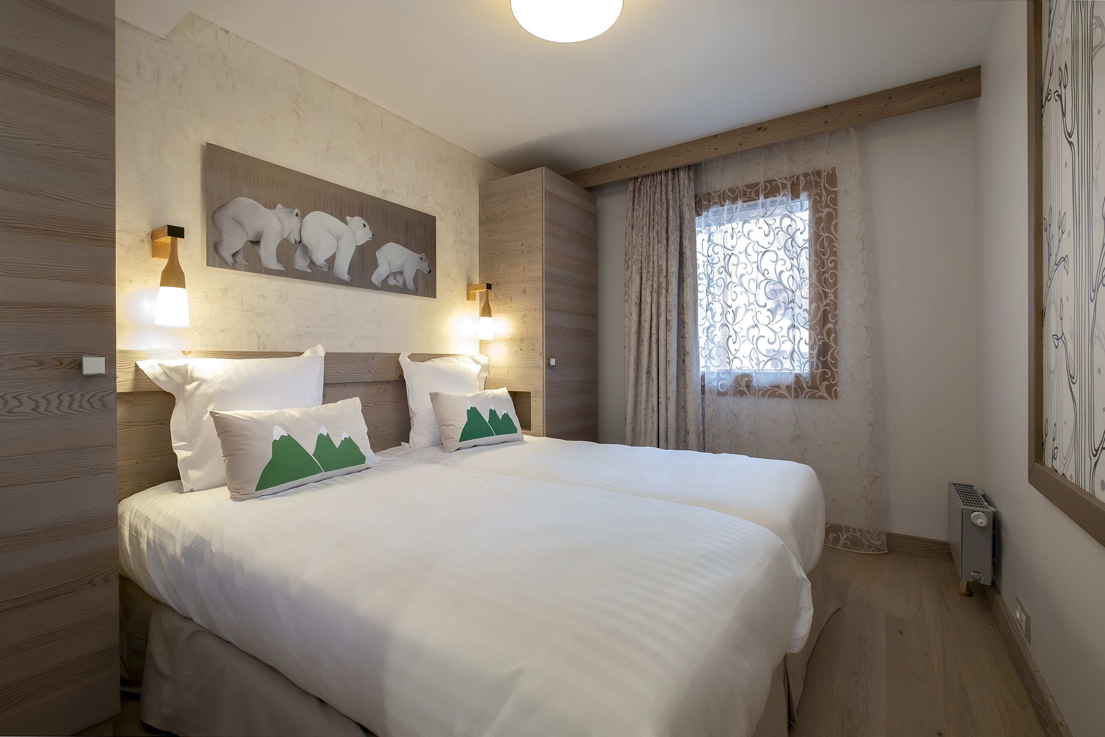 A double bed with white bedding and green mountain pillows, a painting of polar bears on the wall, and a window with patterned curtains.