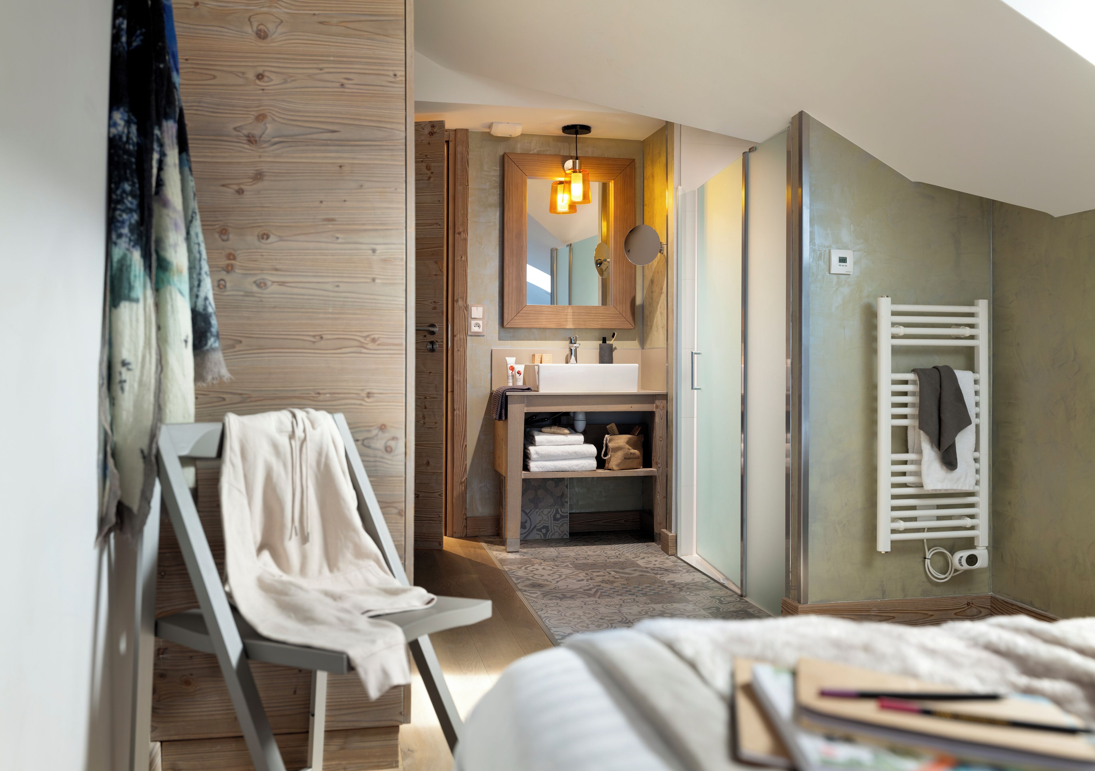 Une chambre d’hôtel moderne avec un plancher en bois, un lit, un bureau avec une chaise, une salle de bain avec un lavabo et un miroir, et un porte-serviettes.