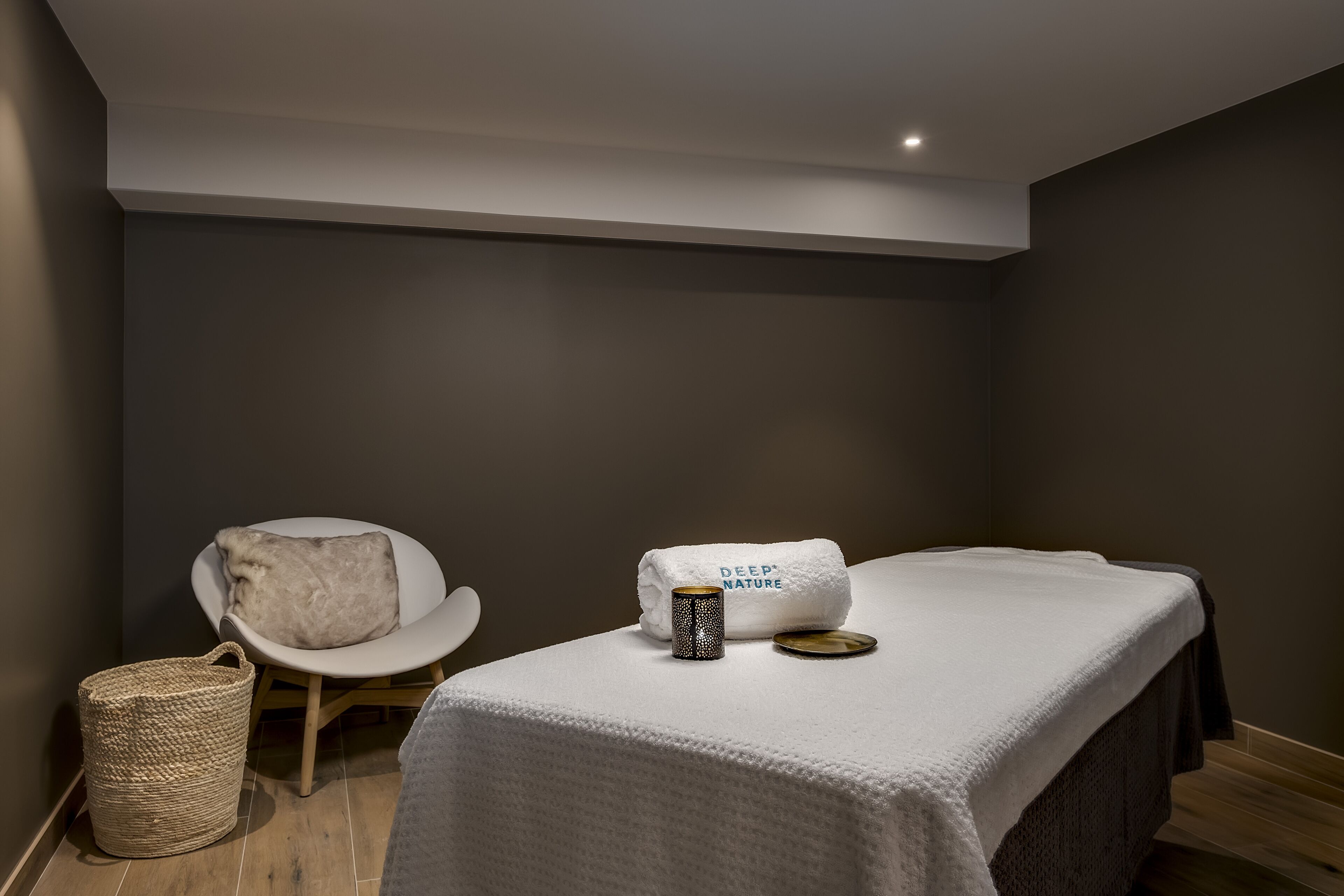Ett spa-rum med en massagebänk, en vit stol, en handduk och en flätad korg.