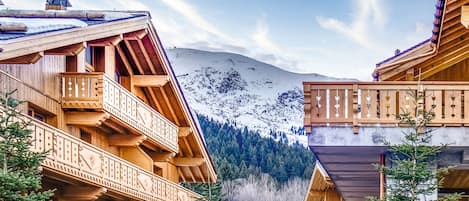 A wooden chalet with balconies, a snowy mountain backdrop, and a forested valley.