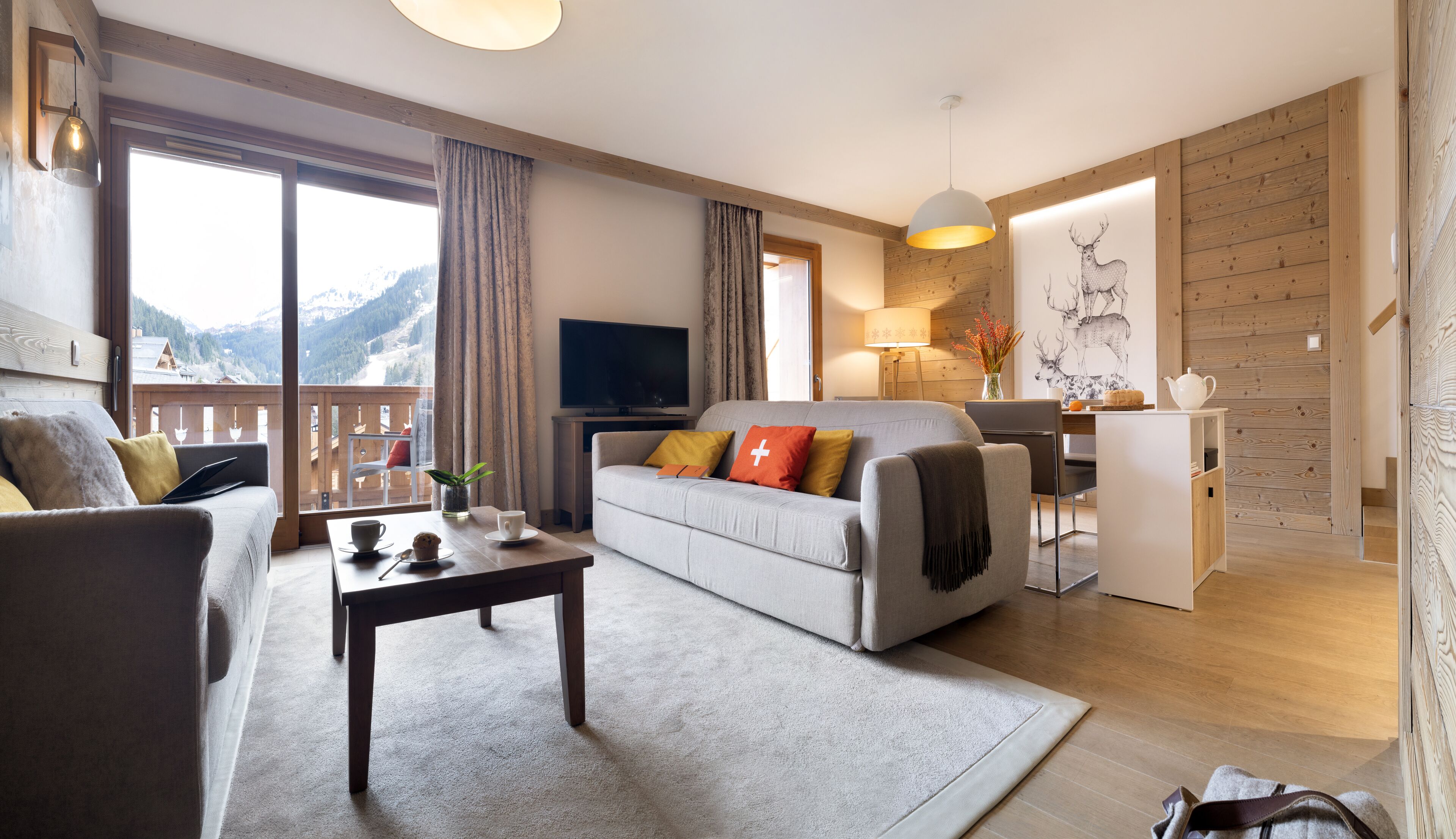 A modern living room with a sofa, coffee table, and a TV. There is a balcony with a view of mountains.