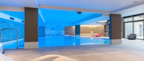 An indoor swimming pool with a clear view of the water, a lounge area with a white chair and a brown cushion, and a stone wall in the background.