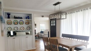 Een eetgedeelte met een houten tafel en stoelen, een wit kastje met decoratieve borden, en een moderne hanglamp.