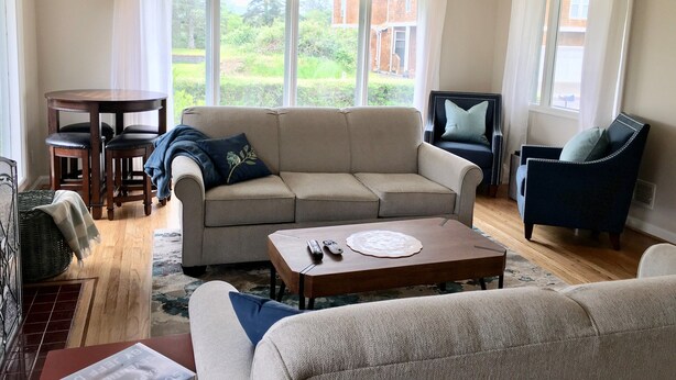 Een woonkamer met een beige bank, een ronde salontafel, een donkerblauwe fauteuil en een klein tafeltje met een afstandsbediening.