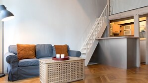 A modern living room with a grey sofa, two brown cushions, a wooden coffee table, and a floor lamp. A staircase leads to an upper level with a kitchen area. - Sunny Dussoubs I Unique 1-BR loft in Montorgueil (Paris)