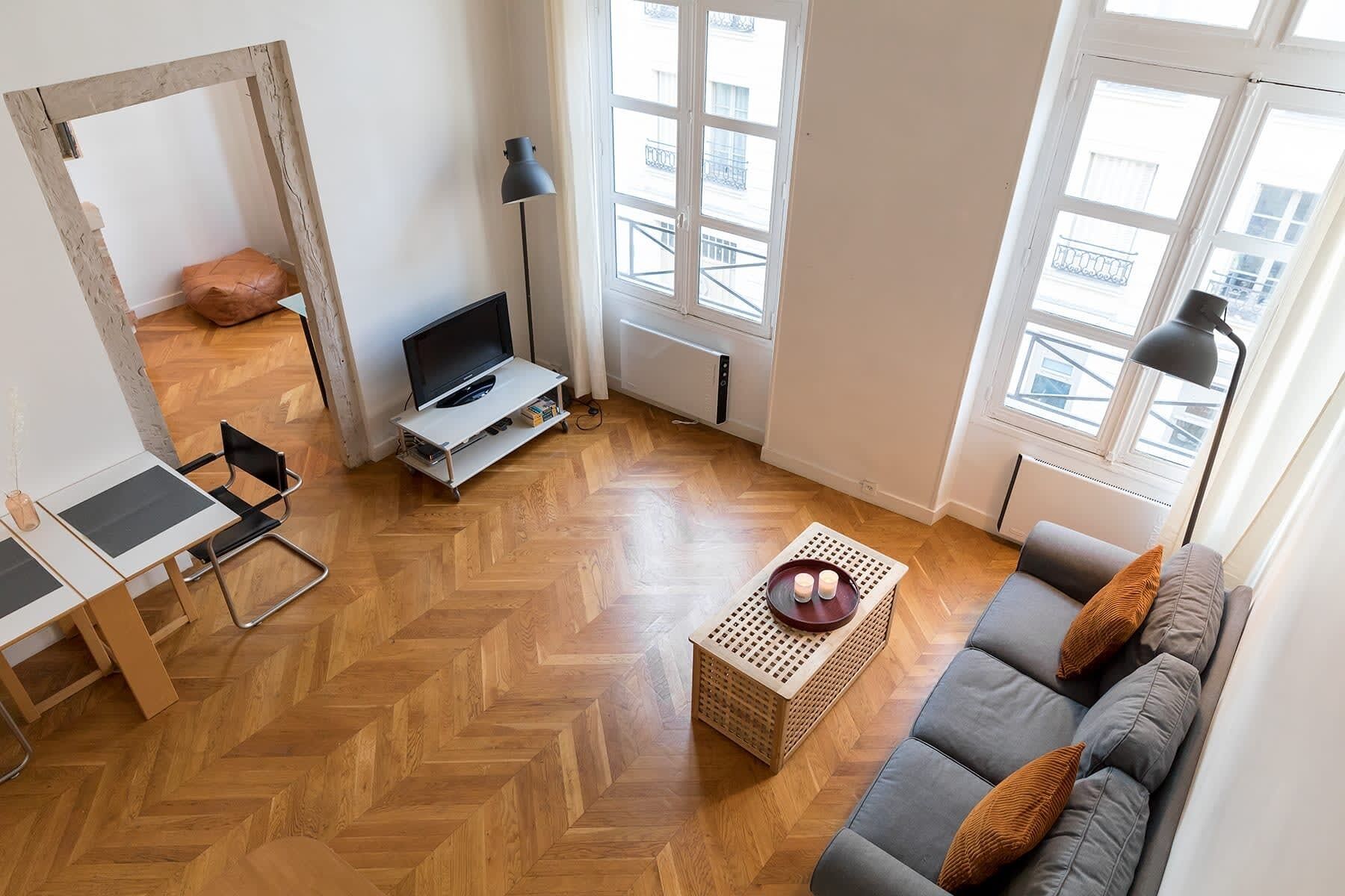 Ein modernes Wohnzimmer mit Fischgrätparkett, einem grauen Sofa mit Kissen, einem kleinen Holztisch, einem Fernseher auf einem Lowboard und einer Lampe.