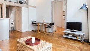 A modern living room with a wooden floor, a TV on a stand, a table with chairs, and a basket with a candle. - Sunny Dussoubs I Unique 1-BR loft in Montorgueil (Paris)