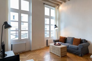 Een moderne woonkamer met een grijze bank, twee zwarte vloerlampen, een houten salontafel en grote ramen met witte kozijnen.