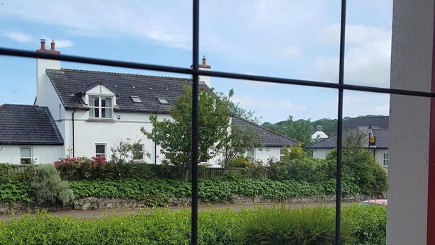 Una veduta attraverso una finestra con un motivo a griglia che mostra una zona residenziale con case, un giardino curato e un vialetto lastricato.