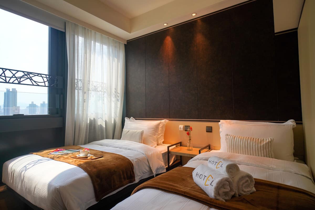 A hotel room with two beds, a city view, a window with curtains, and a nightstand with a vase of flowers.