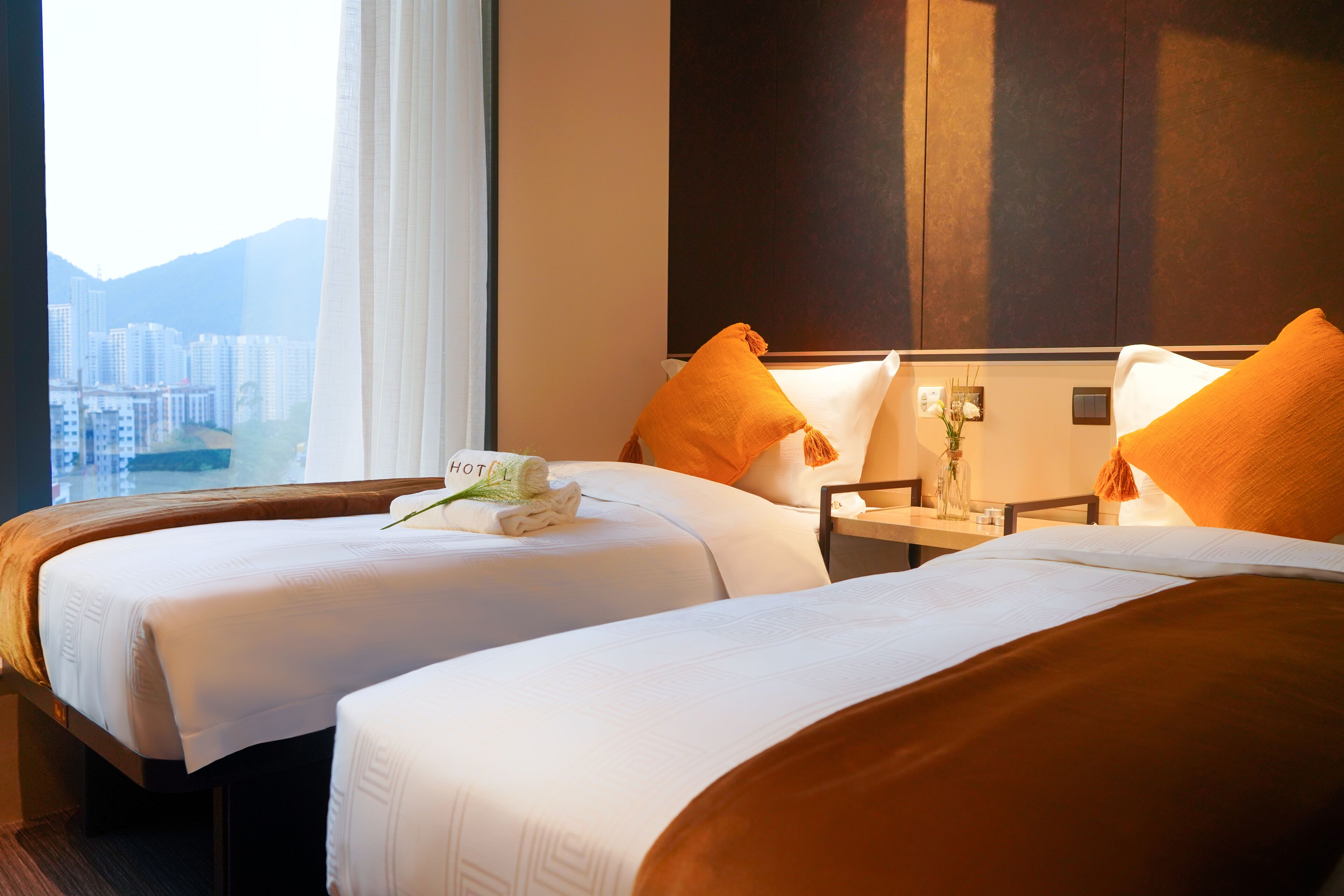 A hotel room with two beds, a nightstand, and a window with a view of buildings.