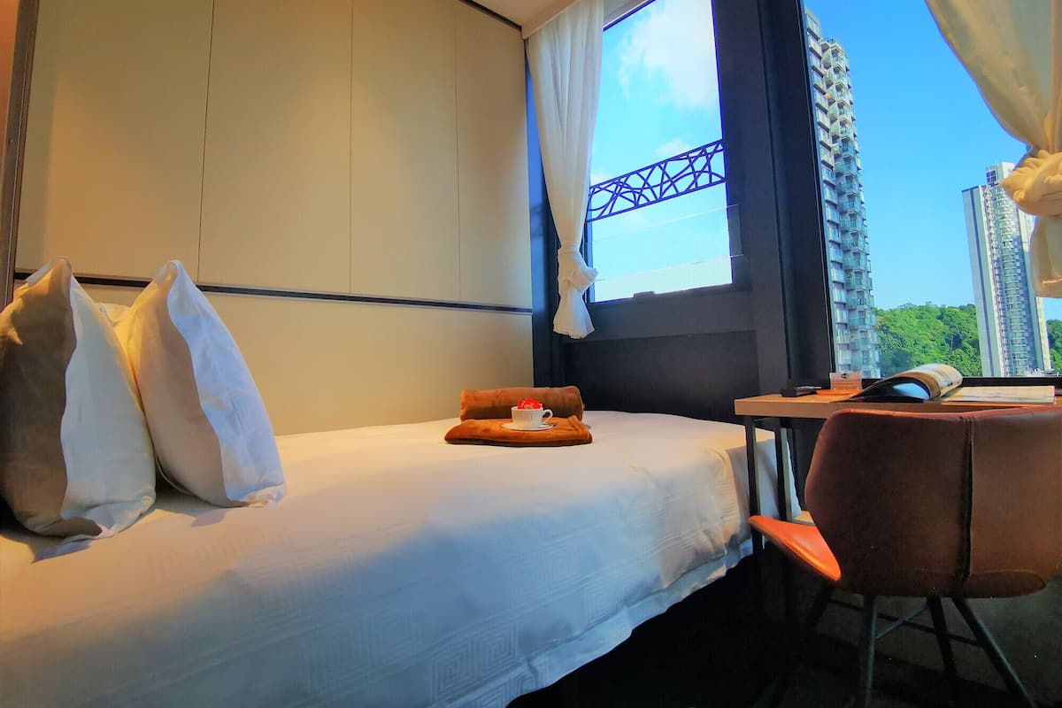 A hotel room with a bed, a small table, and a chair. There is a window with curtains, and a view of tall buildings outside.