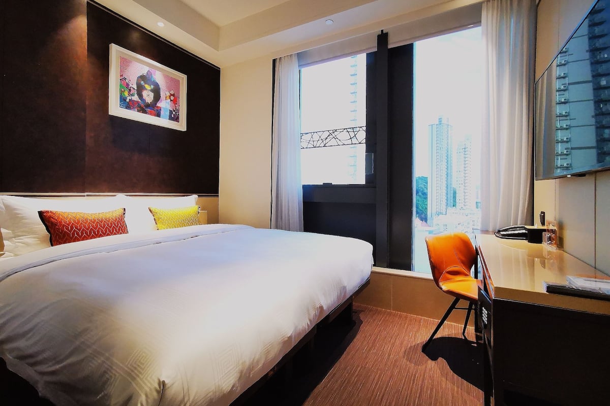 A hotel room with a large bed, a desk, and a chair. There is a window with a view of buildings outside.