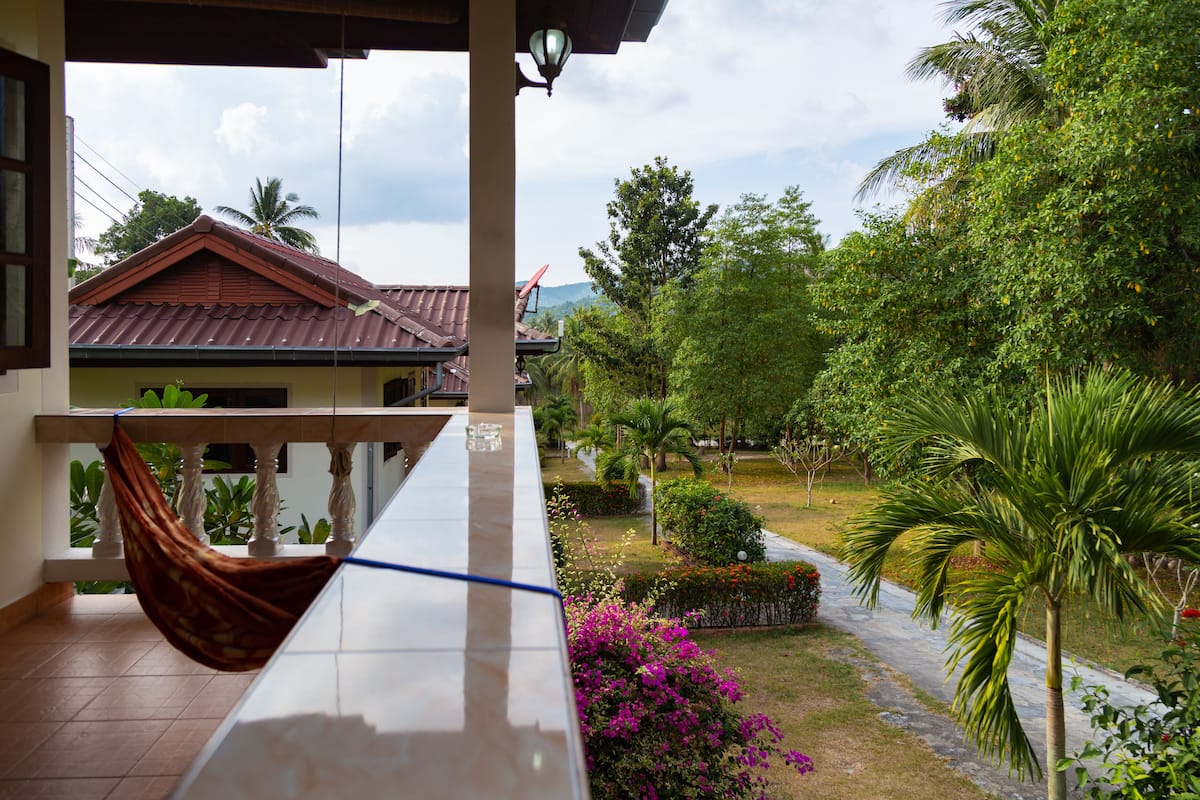 A balcony with a hammock, a view of a garden with palm trees and flowers, and a building with a red roof.