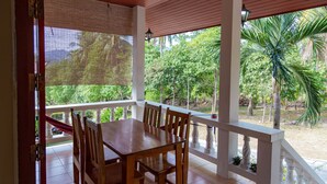 Uma área de refeições ao ar livre com mesa e cadeiras de madeira, vista para vegetação exuberante e piso de azulejos.