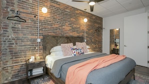 Ein Schlafzimmer mit Backsteinwand, einem Bett mit vielen Kissen, einem Nachttisch, einer Hängelampe und einem Deckenventilator.