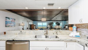 A kitchen with a marble countertop, stainless steel sink, and white cabinetry. There is a view of the ocean through large windows, and a ceiling with wooden beams and recessed lighting. - Seagull 202 - Quiet Beachfront Hideaway with Endless Ocean Views, Swimming Pool, Spa, Washer/Dryer (South Padre Island)