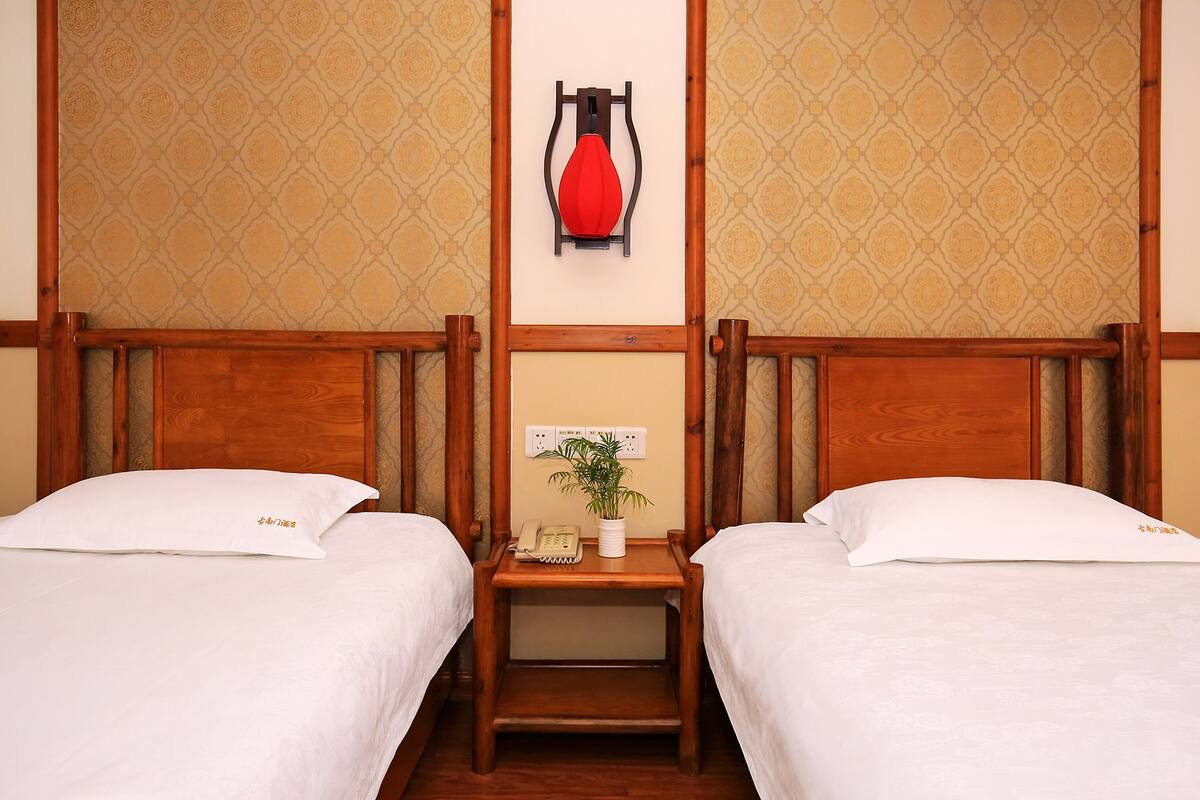 A hotel room with two beds, wooden headboards, a bedside table with a telephone and a plant, and a wall-mounted heater.