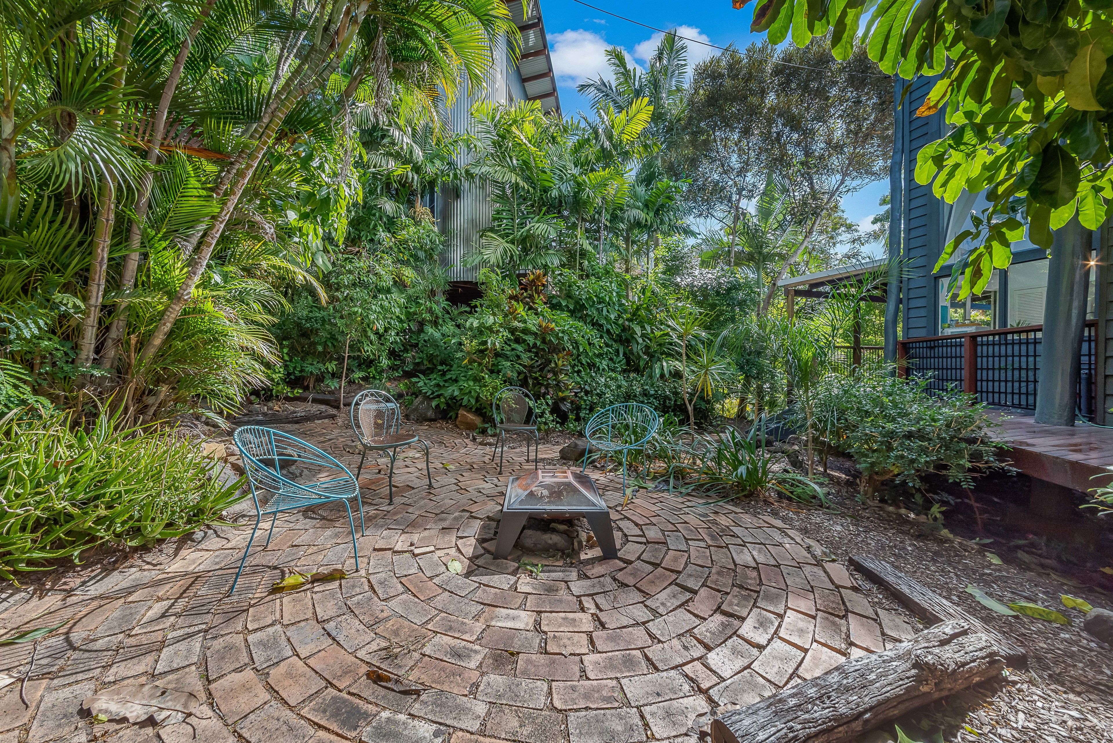 A circular brick patio with metal chairs and a table, surrounded by lush greenery and a building with a blue exterior.