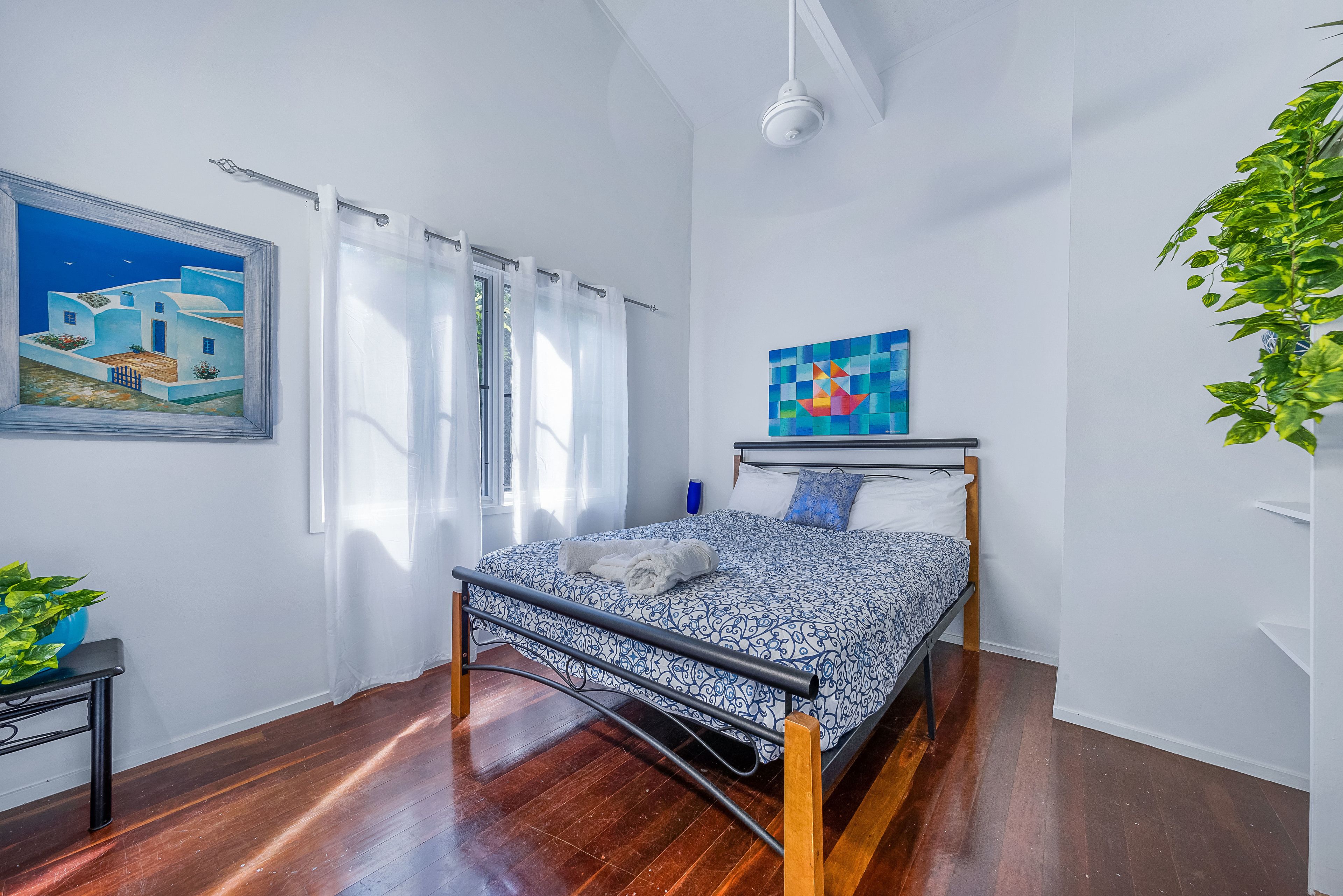 A bedroom with a bed, a window with curtains, a painting on the wall, and a plant.