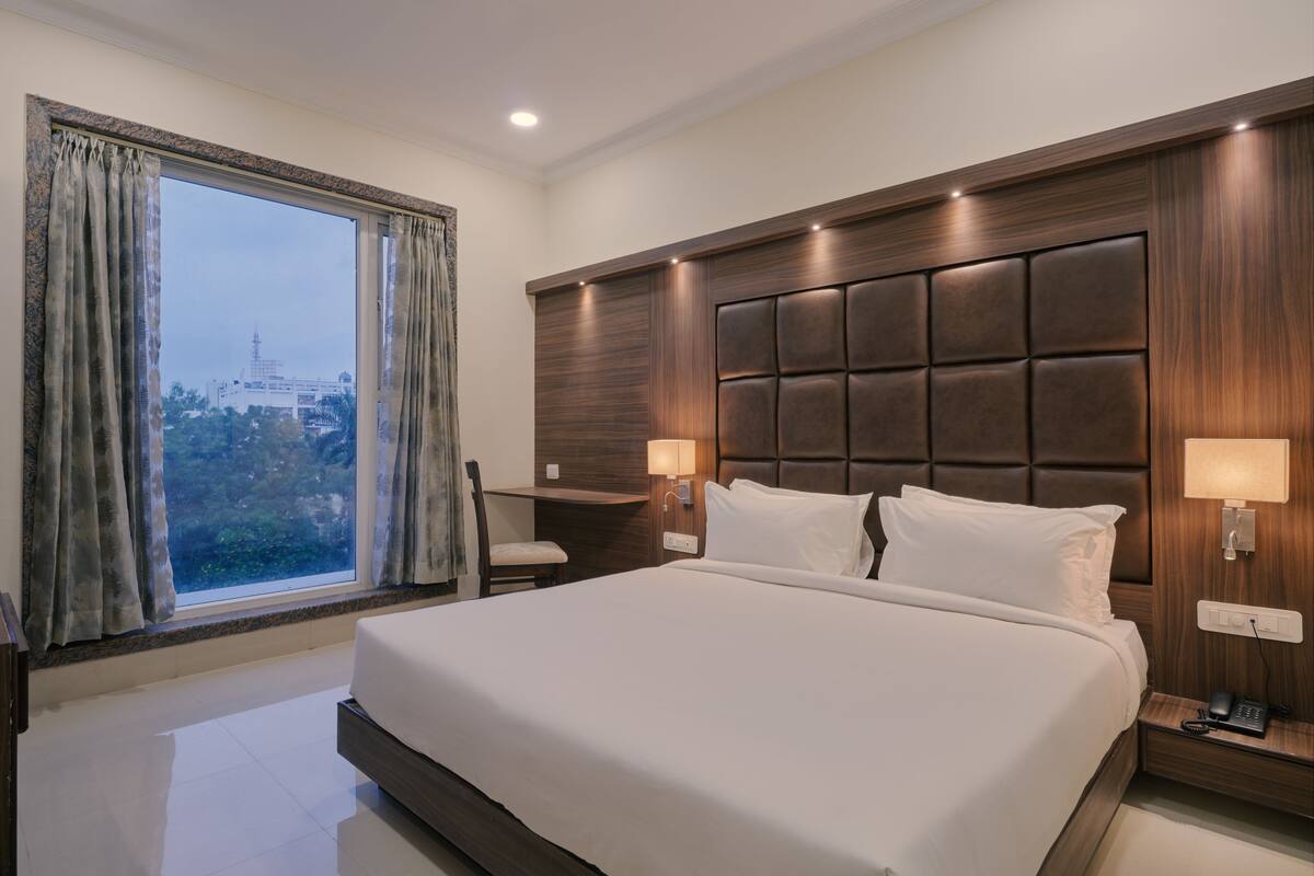 A hotel room with a large bed, a desk, and a view of a cityscape through the window.